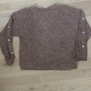 Ljuslila stickad tröja från Lindex Kids - Mysig ljuslila stickad tröja från Lindex Kids med långa ärmar och dekorativa knappar längs ärmarna. Perfekt för kyligare dagar och enkel att matcha med olika outfits.