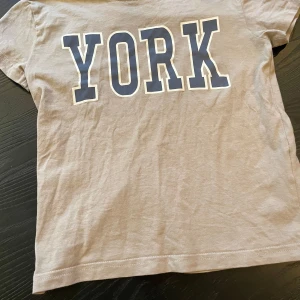 Beige t-shirt med tryck från lager 157 - Säljer en beige t-shirt från lager 157 med stort tryck 'YORK' i mörkblått och vitt på framsidan. T-shirten är i 100% bomull och har en klassisk passform med korta ärmar. Perfekt för en avslappnad stil.