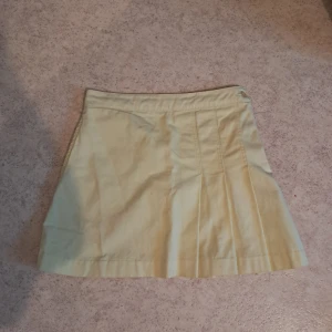 Ljusgul plisserad kortkjol - Säljer en ljusgul kortkjol med plisserade detaljer. Kjolen har dragkedja och knapp i sidan för enkel stängning. Perfekt för dig som gillar en stilren och fräsch look.