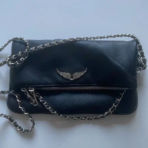 Svart axelväska med kedjerem och vinge-detalj - Säljer en svart axelväska i skinnimitation med silverfärgad kedjerem och dragkedja. Väskan har en snygg vinge-detalj framtill och dragkedjefack både fram och bak. Perfekt för dig som vill ha en stilren accessoar med lite edge. INTE ÄKTA!