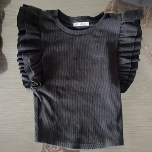 Svart ribbad topp med volangärmar från Zara - Snygg svart ribbad topp från Zara med markerade volangärmar som ger en cool och trendig look. Perfekt att styla till jeans eller kjol för en extra touch. Mjuk och stretchig i materialet.