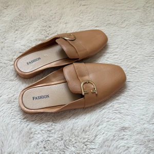 Beige loafers med gulddetalj - Säljer ett par stilrena beige loafers med öppen häl och guldfärgad metallspänne framtill. Skorna har platt sula och är tillverkade i syntetmaterial. Perfekta för en trendig och avslappnad look.