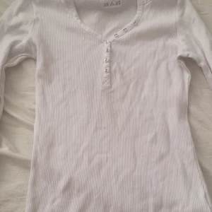 Vit ribbad långärmad topp från Urba Basics - Säljer en vit vintage ribbad långärmad topp från Urban Basics. Tröjan har smala ränder, v-ringning och knappar framtill. Perfekt att styla med jeans eller kjol för en enkel och clean look.