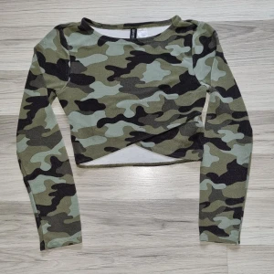 Camouflage croppad långärmad topp från H&M  - Säljer en croppad långärmad topp från H&M i grönt camouflage-mönster. Toppen har en snygg omlott-detalj framtill och är tillverkad i bomullsmix. Perfekt för dig som gillar streetstyle och vill sticka ut.