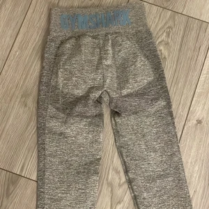 Grå träningstights från Gymshark - Säljer ett par gråmelerade träningstights från Gymshark med hög midja och tight passform. Tightsen har diskret Gymshark-logga i blått på linningen och är tillverkade i ett stretchigt syntetmaterial som passar perfekt till gymmet.
