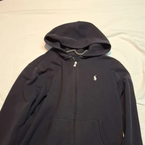 Svart hoodie från Ralph Lauren - Klassisk svart hoodie från Ralph Lauren med dragkedja framtill och vit broderad logga på bröstet. Tröjan har huva och två fickor framtill. Perfekt för en avslappnad stil. Möts i Norrköping 