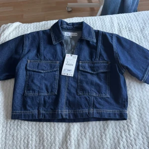 Mörkblå croppad jeansjacka från Zara - Säljer en mörkblå croppad jeansjacka från Zara med två stora bröstfickor och markerade sömmar. Jackan har krage och kort ärm, perfekt för en trendig och avslappnad look.