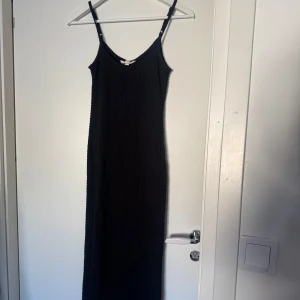 Svart långklänning med smala axelband - En stilren svart långklänning med smala justerbara axelband och rundad halsringning. Klänningen har en figursydd passform. 