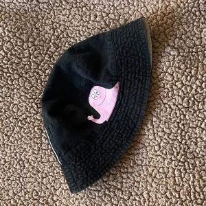 Svart Barbapapa barn bucket hat - Cool svart bucket hat med en rosa Barbapapa-figur broderad framtill. Hatten har en klassisk form och är tillverkad i bomull, perfekt för soliga dagar. Hatten är i barn storlek.