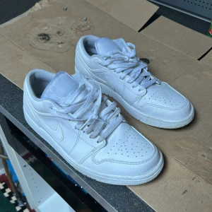 Vita Nike Air Jordans Low - Säljer ett par klassiska vita Nike Air Jordans Low med snörning och perforerad tå. Skorna har en låg siluett och är helt vita med diskreta detaljer och Nike-logga på sidan samt Air Jordan-brodyr på hälen.