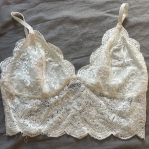 Vit spetsig croppad linnetopp - Säljer en vit croppad linnetopp i spets med blommigt mönster. Toppen har tunna axelband och vacker scallop-kant nertill. Perfekt för att lyfta en outfit med romantisk känsla.