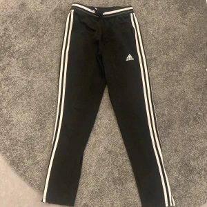 Svarta Adidas träningsbyxor med vita ränder - Klassiska svarta träningsbyxor från Adidas med vita ränder längs sidorna och Adidas-logga på låret. Byxorna har normal passform och resår i midjan. Perfekta för sport eller avslappnade dagar.