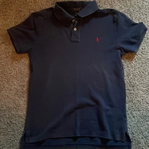 Mörkblå pikétröja från Polo Ralph Lauren - Klassisk mörkblå pikétröja från Polo Ralph Lauren med korta ärmar och krage. Tröjan har två knappar vid halsen och en röd broderad logga på bröstet. Perfekt för en stilren och avslappnad look.