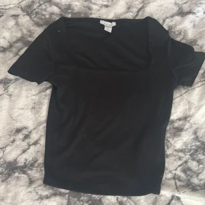 Svart topp med fyrkantig ringning från Amisu - Säljer en svart topp från Amisu med korta ärmar och snygg fyrkantig ringning. Enkel och stilren design som passar perfekt till många olika outfits. Tillverkad i ett mjukt och stretchigt material för en bekväm passform.