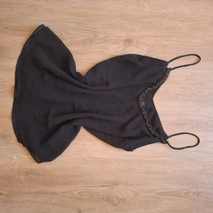 Svart linnetopp med smala axelband och spetsdetalj - Säljer en svart linnetopp med tunna axelband och dekorativ spets längs urringningen. Toppen har en enkel och stilren design, perfekt att matcha med olika outfits. Materialet känns mjukt och följsamt.