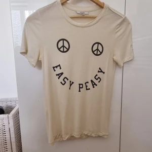 Beige t-shirt med tryck från Ganni - Säljer en beige t-shirt från Ganni med svart tryck framtill som bildar ett smileyface med peace-symboler som ögon och texten 'EASY PEASY' som mun. T-shirten har rund hals och korta ärmar. Perfekt för en chill och avslappnad stil.