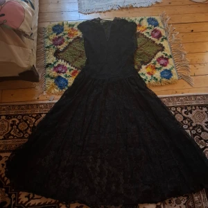 Svart långklänning med spetsdetaljer - Elegant svart långklänning med v-ringning och spetsdetaljer. Klänningen har en klassisk siluett med vid kjol och är ärmlös, perfekt för festliga tillfällen. Materialet ger en lyxig känsla och den svarta färgen gör den tidlös.