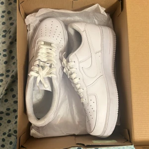 Nike Air Force 1 helvita sneakers - Klassiska helvita Nike Air Force 1 sneakers med perforerad tå, snörning och platt sula. Skorna har en ikonisk design med Nike-logga på sidan och är tillverkade i syntetmaterial och skinn.
