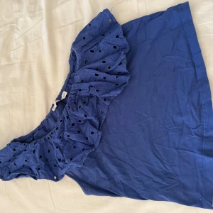 Blå ärmlös topp med volang från Mango - Säljer en blå ärmlös topp från Mango med bred volang längs ena axeln. Toppen har ett snyggt hålmönster i volangen och är tillverkad i ett mjukt material. Perfekt för varma dagar!