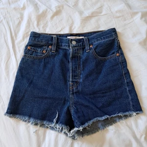 Levi's blå jeansshorts med fransar - Säljer ett par klassiska blå jeansshorts från Levi's med hög midja och råa franskantade benslut. Shortsen har fem fickor, knappgylf och tydliga sömmar. Perfekta för sommaren!