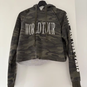 Camouflage hoodie från Gina Tricot - Säljer en croppad hoodie från Gina Tricot i camouflage-mönster med texten 'WORLD TOUR' framtill och städer på ärmen. Tröjan har huva, långa ärmar och är i mjukt material. Perfekt för en avslappnad streetstyle-look. Nopprig 