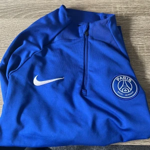 Blå Paris Saint-Germain träningströja från Nike - Säljer en blå långärmad träningströja från Nike med Paris Saint-Germain-logga på bröstet. Tröjan har halv dragkedja framtill och är perfekt för fotbollsträning. Klassisk Nike Swoosh på ena sidan av bröstet.