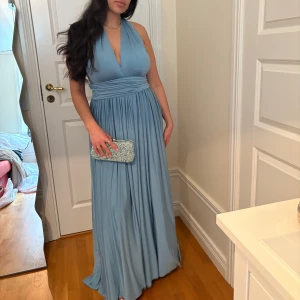 Festklänning - Ljusblå långklänning - Säljer en elegant ljusblå långklänning med djup v-ringning. Klänningen går att ha i olika modeller genom att kunna knyta den på olika sätt! Klänningen är helt oanvänd