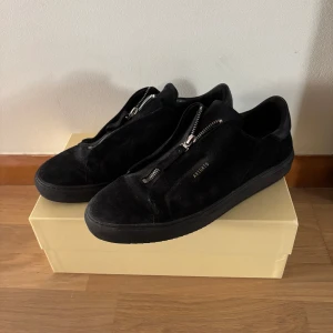 Svarta sneakers från Axel Arigato med dragkedja - Säljer ett par svarta sneakers från Axel Arigato i mocka med dragkedja på ovansidan istället för klassisk snörning. Skorna är knappt använda, skick 9.5/10. Storlek 44 