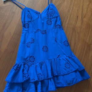 Blå kortklänning med broderade blommor - Säljer en blå kortklänning med smala axelband och volangkant nertill. Klänningen har broderade blommor och en lätt v-ringning. Perfekt för varma dagar och sommarens alla tillfällen.