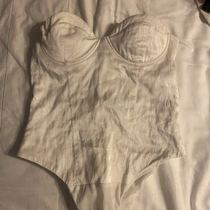 Vit bandoue body med kupa - Säljer en axelbandslös body i skimmer vit färg. Med markerade kupor/bygel som skapar en bra siluett.