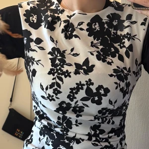 Vit och svart blommig tight klänning - Säljer en vit klänning med svart blommigt mönster. Modellen är tight och ärmlös med rund halsringning. Perfekt för dig som gillar en klassisk men ändå trendig look. Materialet känns mjukt och stretchigt, vilket gör den bekväm att bära.