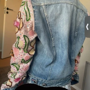 Jeansjacka med broderade ärmar från Rinascimento - Unik jeansjacka från Replay med ljusblå denim och detaljerade broderade ärmar i rosa, grönt och gult. Ärmarna är dekorerade med pärlor, paljetter och blommönster för en färgstark look. Klassisk passform med knappar framtill.