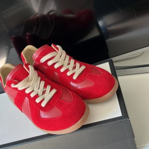Röda sneakers från Maison Margiela  - Säljer ett par Maison Margiela sneakers i rött med beige snörning och gummisula. Skorna har en mix av mocka och skinn, klassisk siluett och rund tå. Perfekta för dig som vill sticka ut med färg på fötterna. Och är riktigt fetta och dom är helt nya, jag råkade köpa fel storlek så det är därför jag säljer. Nu när sommarn kommer är det här dom snyggaste skorna du kan ha.