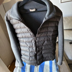 Grå pufferjacka från Moncler - Snygg grå cardigan från Moncler med quiltad framsida och ärmar i stickad bomull. Jackan har dragkedja framtill, hög krage. Perfekt för kyliga sommardagar. Använd ett fåtal gånger 