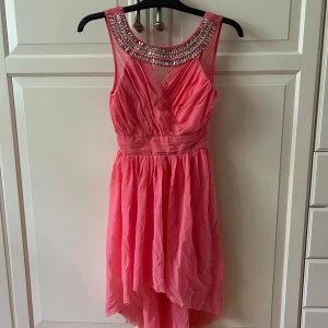 Rosa festklänning med strass från Elise Ryan - Supersnygg rosa klänning från Elise Ryan med glittrande strassdetaljer runt halsringningen. Klänningen har omlottliv, markerad midja. Tyget är lätt och faller fint, perfekt för festliga tillfällen. I strl Xs
