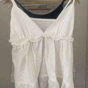 Vit linnetopp med spetsdetaljer - Söt vit linnetopp med smala axelband och v-ringning. Toppen har volang och spetsdetaljer längs bröstet och nederkanten samt smock i ryggen för extra komfort. Perfekt för varma dagar. Banden är justerbara och pris kan diskuteras.  Vet inte vart de är ifrån då de inte finns någon lapp men det är storlek xs. Aldrig använd 