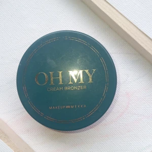 OH MY Cream Bronzer från Makeup Mekka - Krämig bronzer i burk från Makeup Mekka i nyansen 'Oh My Bronze'. Ger en naturlig solkysst look med en mjuk, brun ton. Kommer i en rund, grön förpackning med guldtext och innehåller 5 g produkt.