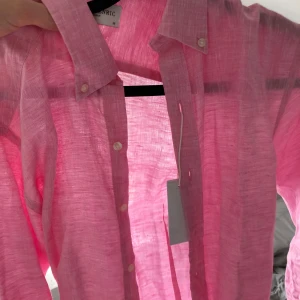 Rosa linneskjorta från Henric - Säljer en snygg rosa skjorta i linne från Henric. Skjortan har klassisk krage, vita knappar och lång ärm. Perfekt för dig som vill ha en luftig och stilren look i sommarvärmen.