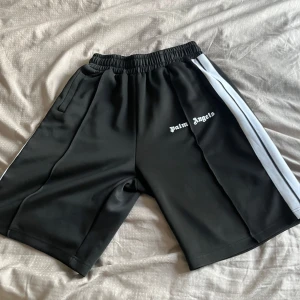 Palm Angels Shorts - Svarta shorts med vit sidoremsa och elastisk midja från Palm Angels. Shortsen har fickor på sidorna och logotypen tryckt på vänster ben. Perfekta för en sportig och avslappnad stil. Storlek S men passar även M
