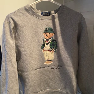 Grå sweatshirt med Polo Bear från Ralph Lauren - Grå sweatshirt från Ralph Lauren med ikoniska Polo Bear-trycket framtill. Tröjan har rund halsringning och långa ärmar. Klassisk och stilren design med mjukt bomullsmaterial, perfekt för en avslappnad look. Storlek 160 cm