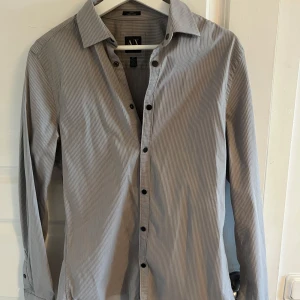 Gråvit randig skjorta från Armani  - Säljer en klassisk gråvit randig skjorta från Zara Man. Skjortan har lång ärm, svarta knappar och en smal passform. Perfekt för dig som gillar stilrena och enkla plagg med diskret mönster. Aldrig använd