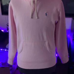Polo raulph lauren hoodie - Säljer en ljusrosa hoodie från Polo Ralph Lauren med klassisk broderad logga på bröstet. Tröjan har huva med dragsko och en stor magficka. Perfekt för dig som gillar stilrena och bekväma plagg. Säljer för 1000 kr då den kostar c:a 2000 kr annars. Har inte använt den så många gånger. Säljer pågrund av att jag tröttnat på denna tröja.
