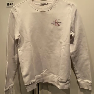 Vit sweatshirt från Calvin Klein Jeans - Säljer en stilren vit sweatshirt från Calvin Klein Jeans med diskret CK-logga i rosa och svart på bröstet. Tröjan har rund halsringning och långa ärmar. Perfekt för en clean och enkel look.