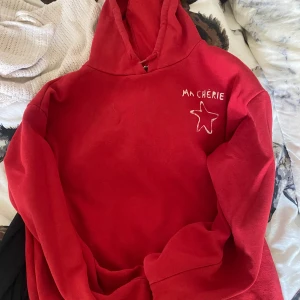 Röd hoodie med tryck från Gina Tricot - Röd hoodie från Gina Tricot med vit text 'MA CHÉRIE' och en stjärna tryckt på bröstet. Tröjan har huva och långa ärmar, samt ribbade muddar vid ärmslut och nederkant. Perfekt för dig som gillar en chill och färgstark stil.