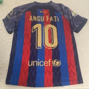 FC Barcelona Ansu Fati #10 tröja - Snygg FC Barcelona fotbollströja med Ansu Fati och nummer 10 på ryggen. Tröjan har klassiska blå och röda ränder, korta ärmar och tryckta detaljer i guld. Materialet är lätt och andas, perfekt för match eller träning.