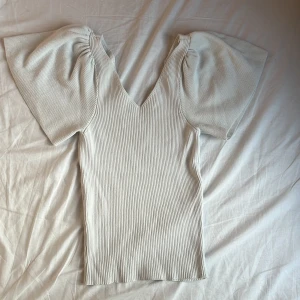 Vit ribbad topp med puffärm från ONLY - Supersnygg ribbad topp i färgen eggnog från ONLY Petite. Toppen har v-ringning, korta puffärmar och en figurnära passform. Perfekt för dig som gillar en stilren look med lite extra volym på axlarna. Materialet är mjukt och stretchigt för skön känsla. Färgen på tröjan är lite urtvättad och banden man ska knyta i ryggen finns inte kvar, Nypris 329 kr.