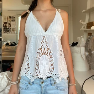 Vit spetsig linnetopp med broderi - Superfin vit linnetopp med broderade spetsdetaljer och v-ringning. Toppen har tunna axelband som korsas i ryggen och en luftig, flowy passform. Perfekt för varma dagar och ger en romantisk vibe med sina genombrutna spetsmönster och dekorativa kanter.