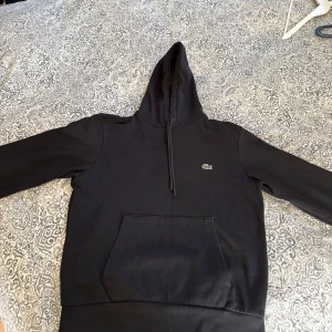 Svart hoodie från Lacoste med huva - Svart hoodie från Lacoste med klassisk krokodillogga på bröstet. Tröjan har huva med snörning, stor magficka och långa ärmar. Perfekt för en chill och stilren look.