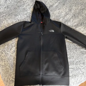 Svart zip up från The North Face - Svart zip up med huva från The North Face. Tröjan har dragkedja framtill, två stora fickor och logga på bröstet. Tillverkad i ett slätt syntetmaterial som känns sportigt och stilrent. Perfekt för dig som gillar enkel och clean look.