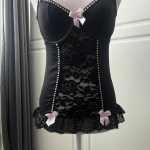 Svart korsettopp med spets och rosetter - Supersnygg svart korsettopp med blommig spets framtill, volangkant nertill och söta ljusrosa rosetter. Storlek strl M men passar även S